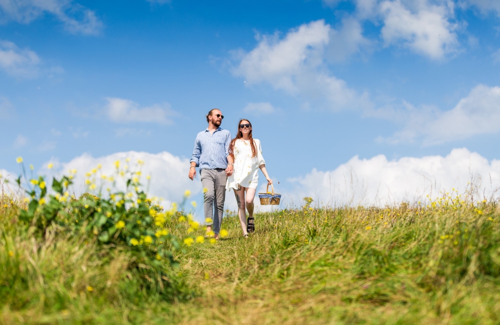 a smiling couple walking through the grassy downs with a picnic basket