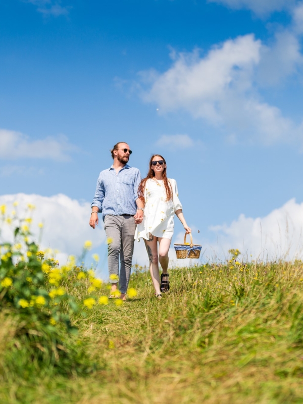 a smiling couple walking through the grassy downs with a picnic basket