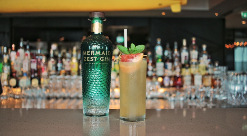 a green glass bottle of mermaid zest gin beside a glass containing a yellow coloured cocktail and fruit garnish