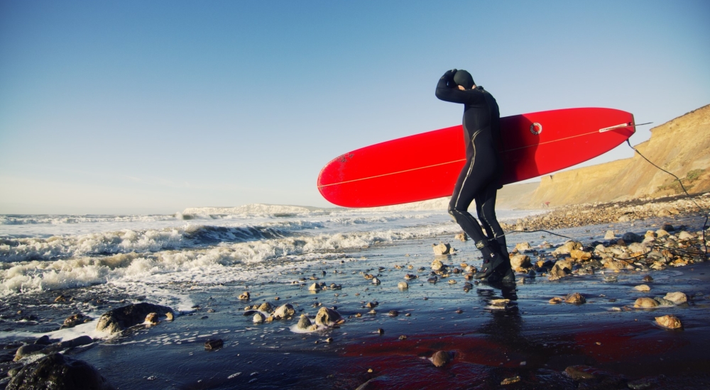 a surfer on the beach in a black wetsuit, holding a red surfboard